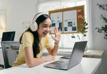 Poster - Young businesswoman is waving while making a video call on her laptop. She is wearing headphones and smiling at the camera