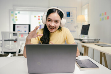 Poster - Young asian woman is making a video call on her laptop and gesturing with her hands while sitting at her desk in her startup's bright office