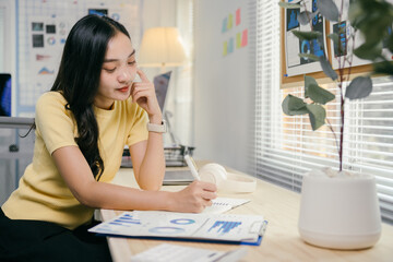 Aufkleber - Young asian woman focused on analyzing charts and jotting notes in her cozy home office, planning and strategizing for her business