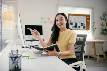 Poster - Asian businesswoman is sitting at her desk and holding a notebook while pointing with a pen at a computer screen. She is smiling and appears to be happy with her work