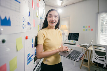 Poster - Young professional is leaning against a whiteboard filled with charts and graphs, holding her laptop and smiling while working on a marketing strategy