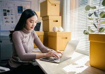 Aufkleber - Young asian woman is working on a laptop in her home office surrounded by cardboard boxes, managing her online business