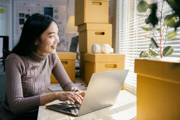 Poster - Young entrepreneur working on a laptop, surrounded by cardboard boxes, in her home office, illuminated by natural light. She is smiling and looking away from the camera