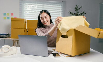 Aufkleber - Young asian woman entrepreneur happily packing clothes in a cardboard box at her home office. Managing her startup business with a laptop for smooth delivery and excellent customer service