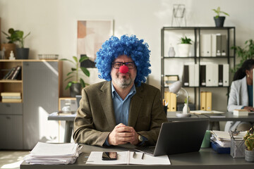 Poster - Serious mature male boss in clown wig looking at you while sitting by workplace with laptop and paper documents