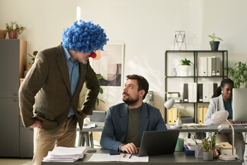 Poster - Young businessman looking at mature male boss in clown wig giving him working instructions while standing in front of subordinate