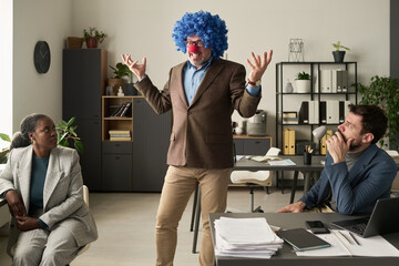 Poster - Annoyed mature clown boss standing between two young intercultural tense subordinates sitting by their workplaces