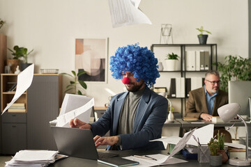 Poster - Young serious male economist in clown wig typing on laptop keypad while several paper documents falling on his workplace