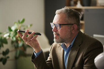 Poster - Mature male boss in elegant suit recording voice message while sitting in front of camera and holding smartphone by his mouth