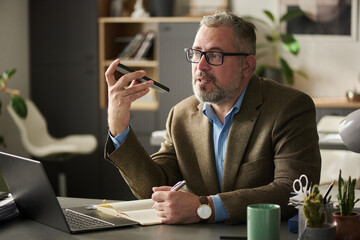 Poster - Confident male broker in eyeglasses and suit sitting by workplace in office and recording voice message for subordinate