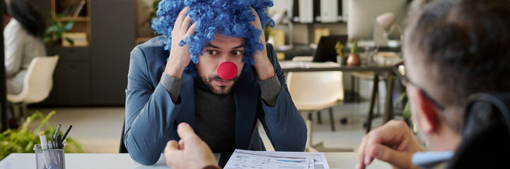 Poster - Header of young stressed businessman in clown wig and nose sitting in front of boss and holding head in hands during discussion