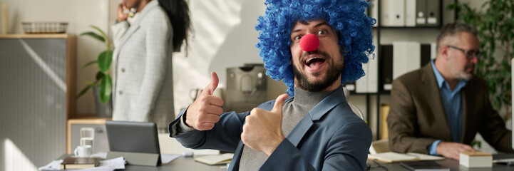 Poster - Header of young successful office worker in clown wig and nose looking at camera and showing thumbs up while sitting by workplace