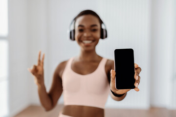 Aufkleber - Sports Application. Excited fit young sporty lady in headset holding and showing smartphone with empty screen for mockup template making peace sign v victory gesture, blurred background selective