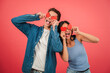 © Jose Calsina - Playful young couple holding red heart shapes and making funny faces against a vibrant red background, celebrating love and happiness with joyful expressions in a fun and creative Valentine