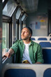 © DimaBerlin - Thoughtful man riding public transportation, looks out window with interest, enjoys calm trip with music in headphones using phone. Focused male tourist travel by electric train reflection about life
