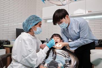 Wall Mural - Asian female pediatric dentist examines a girl's teeth in dental clinic and takes care and encourages of father, mouth oral hygiene, and professional orthodontic healthcare work in a kid hospital.