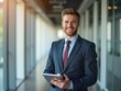 © NETI studio - Confident young professional dressed smartly, smiling and holding a tablet, standing in a modern office hallway with natural lighting, representing leadership and innovation in a business environment
