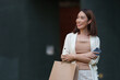 © crizzystudio - Smiling asian woman holding a shopping bag and smartphone, looking away while enjoying a leisurely day of shopping in an urban outdoor setting, radiating happiness and confidence