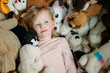 © Cavan Images - Child lying amidst a collection of stuffed animals, looking content.