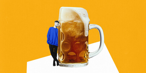 Man wearing blue sweatshirt leaning against oversized beer mug with foam spilling over rim against bright yellow background. Creative art. Concept of beer, party, celebration, festival