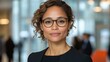 © imagineRbc - A woman with curly hair, wearing glasses, stands in a bustling office filled with natural light. She appears confident and ready for a meeting, embodying professionalism