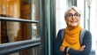 © imagineRbc - Older woman with gray hair and glasses, wearing a yellow scarf, stands by a shop window in a vibrant urban area. She appears content and relaxed, enjoying the sunny day