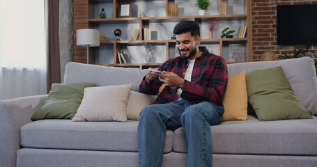 Attractive indian man using smartphone recording answering by audio message in social network while sitting on couch in light living room. Communication, internet.