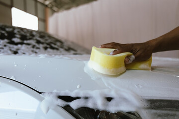 A professional car cleaner meticulously washing a shiny vehicle using a pressure hose and foam. The man focuses on the car's exterior, ensuring a spotless, polished finish at an auto detailing station