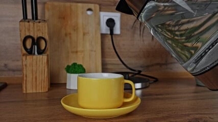 Wall Mural - A yellow mug with a saucer into which hot water for tea is poured from a kettle, they stand on a wooden table, behind it you can see plants and wooden kitchen utensils. Concept of tea drinking, 5