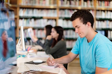 Poster - Young man in a blue shirt writing at a desk in a library. Focused teen student take notes in library with other study in the background. Young man student and other studying with computer in library.