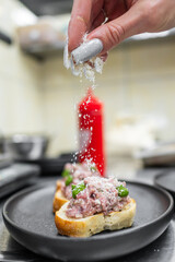 Wall Mural - A close-up of a hand sprinkling grated cheese over a gourmet bruschetta topped with a savory mixture, served on a black plate. Bright kitchen background adds to the culinary atmosphere.