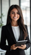 © Michael - Young businesswoman wearing a stylish suit, holding a tablet and smiling confidently in a modern office environment