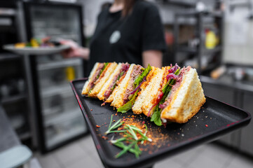 Wall Mural - A close-up of a neatly arranged sandwich platter on a black tray, featuring layers of meat, greens, and herbs. A person partially visible in the background, enhancing the culinary setting.