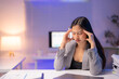 © Tj - Young Asian businesswoman experiencing a headache while touching her temples, working late at night with a laptop and financial reports in a busy office environment