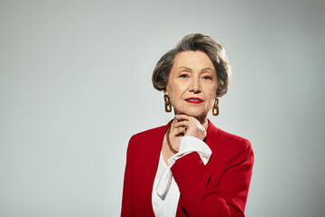 A stylish woman in her 60s radiates grace in elegant red against a grey backdrop.