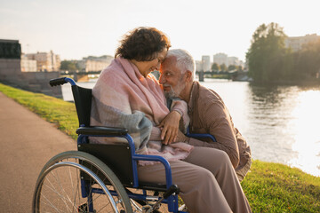 Wall Mural - Senior woman in wheelchair walking with caregiver old man on road in park. Elderly family couple man supporting embracing woman in chair for people with disability outdoor. Rehabilitation