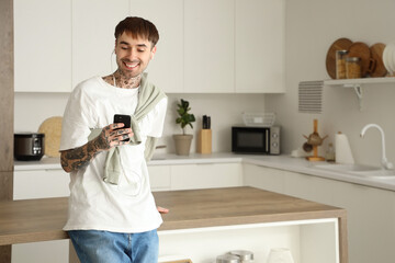 Wall Mural - Young tattooed man with earphones and mobile phone listening to music in kitchen