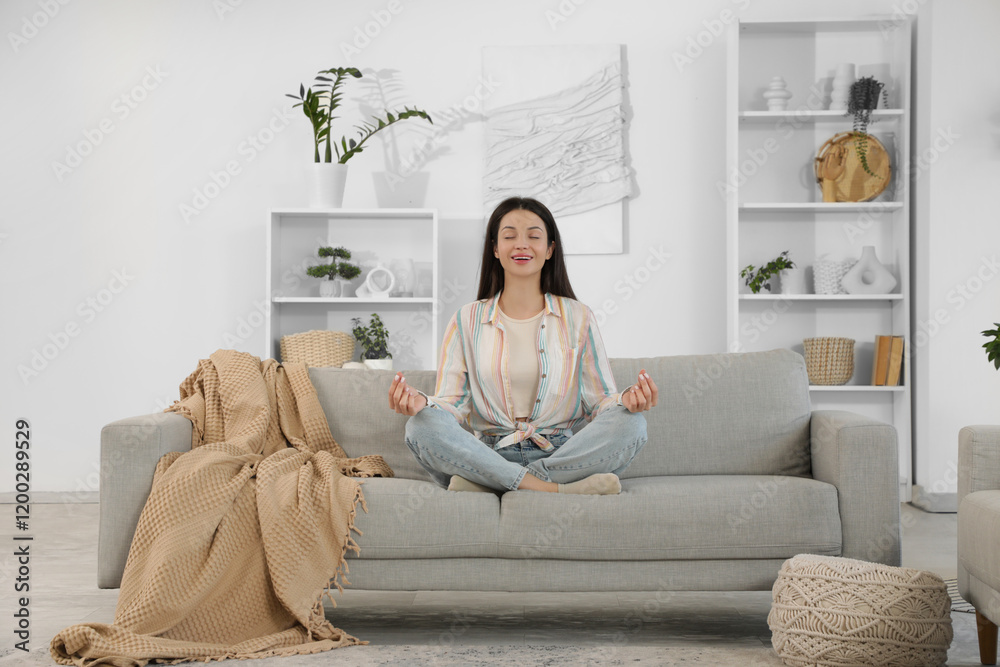 Beautiful young woman meditating on cozy sofa at home