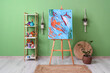 © Pixel-Shot - Painting on easel and art supplies in shelving units near green wall
