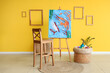 © Pixel-Shot - Easel with painting, chair and art supplies on wicker table near yellow wall