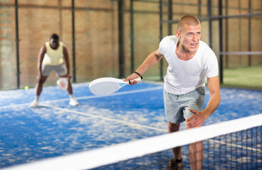 Wall Mural - Portrait of emotional paddle tennis player swinging white racket, ready to hit ball on indoor court