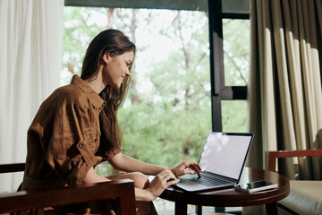 Poster - Young woman working on a laptop in a cozy hotel room, showcasing a focused expression, dressed in a casual brown outfit, surrounded by greenery