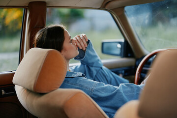 Canvas Print - Young woman resting in a vintage car, wearing a denim jacket, looking thoughtful, with a serene expression and natural background, evoking nostalgia and calmness.