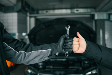 Poster - Close-up of a mechanic's gloved hand holding a wrench and a customer's hand giving a thumbs-up, symbolizing satisfaction and trust in car repair services