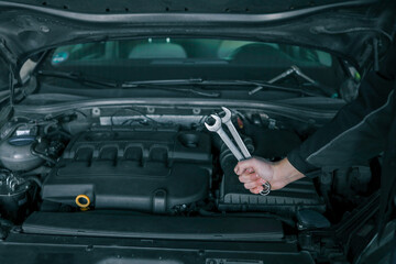 Fototapete - A mechanic's hand holding two wrenches over an open car engine, showcasing tools used for vehicle maintenance. The car's hood is open, revealing the engine components