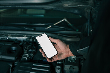 Aufkleber - Mechanic holding a smartphone with a blank white screen in front of an open car engine for diagnostics or maintenance. Indoor workshop setting