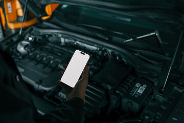 Aufkleber - Mechanic holding a smartphone with a blank white screen in front of an open car engine for diagnostics or maintenance. Indoor workshop setting