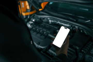 Aufkleber - Mechanic holding a smartphone with a blank white screen in front of an open car engine for diagnostics or maintenance. Indoor workshop setting