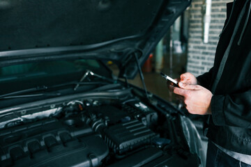 Aufkleber - Mechanic using a smartphone while inspecting an engine under the open hood of a car in a professional garage. Modern technology in automotive diagnostics and repair