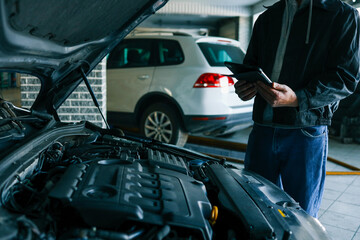 Aufkleber - A mechanic holding a tablet while inspecting a car engine in a workshop. The focus is on the integration of modern technology in vehicle diagnostics and repair